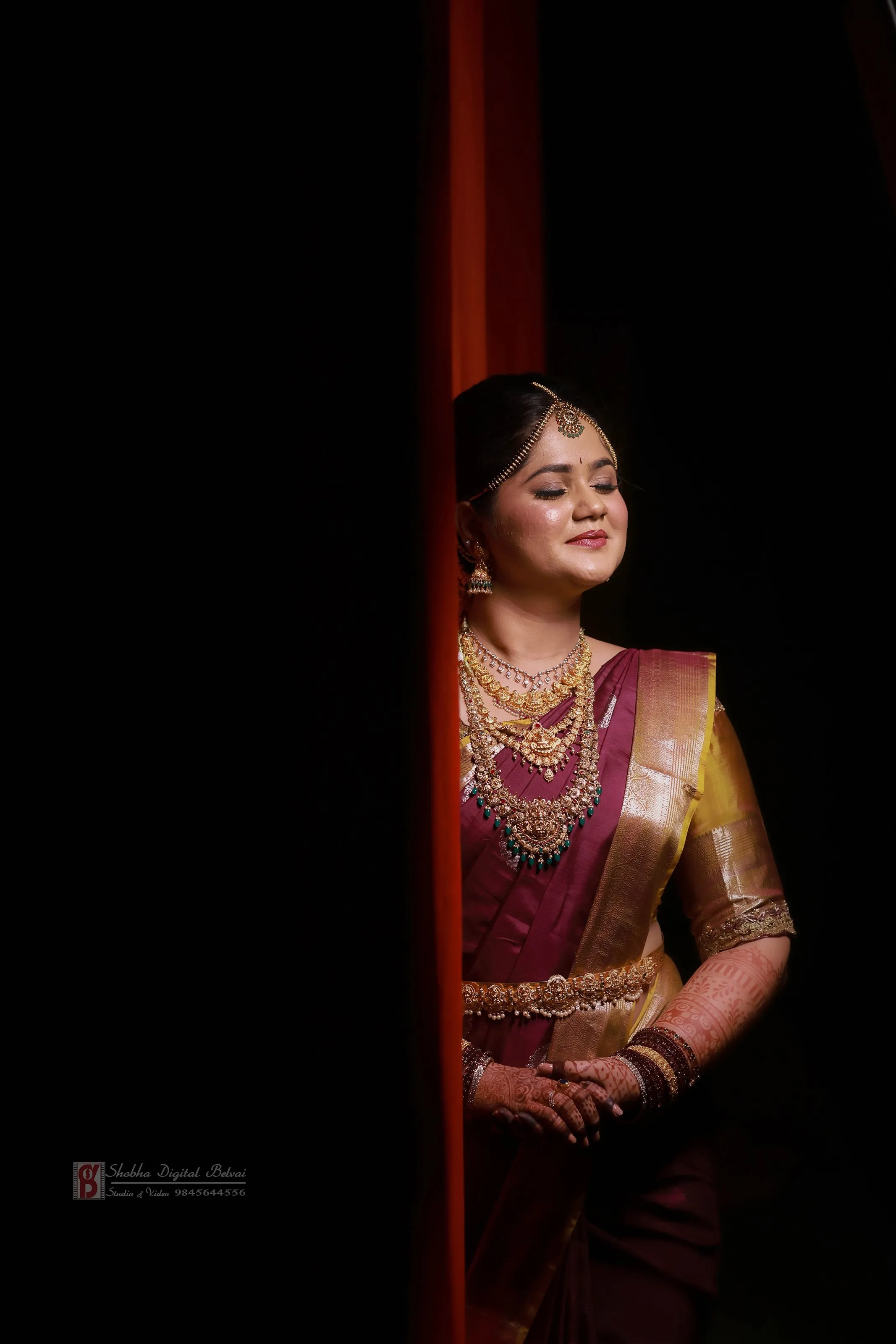 A bride in a maroon and gold silk saree with traditional temple jewellery stands with eyes closed, softly lit beside an orange curtain