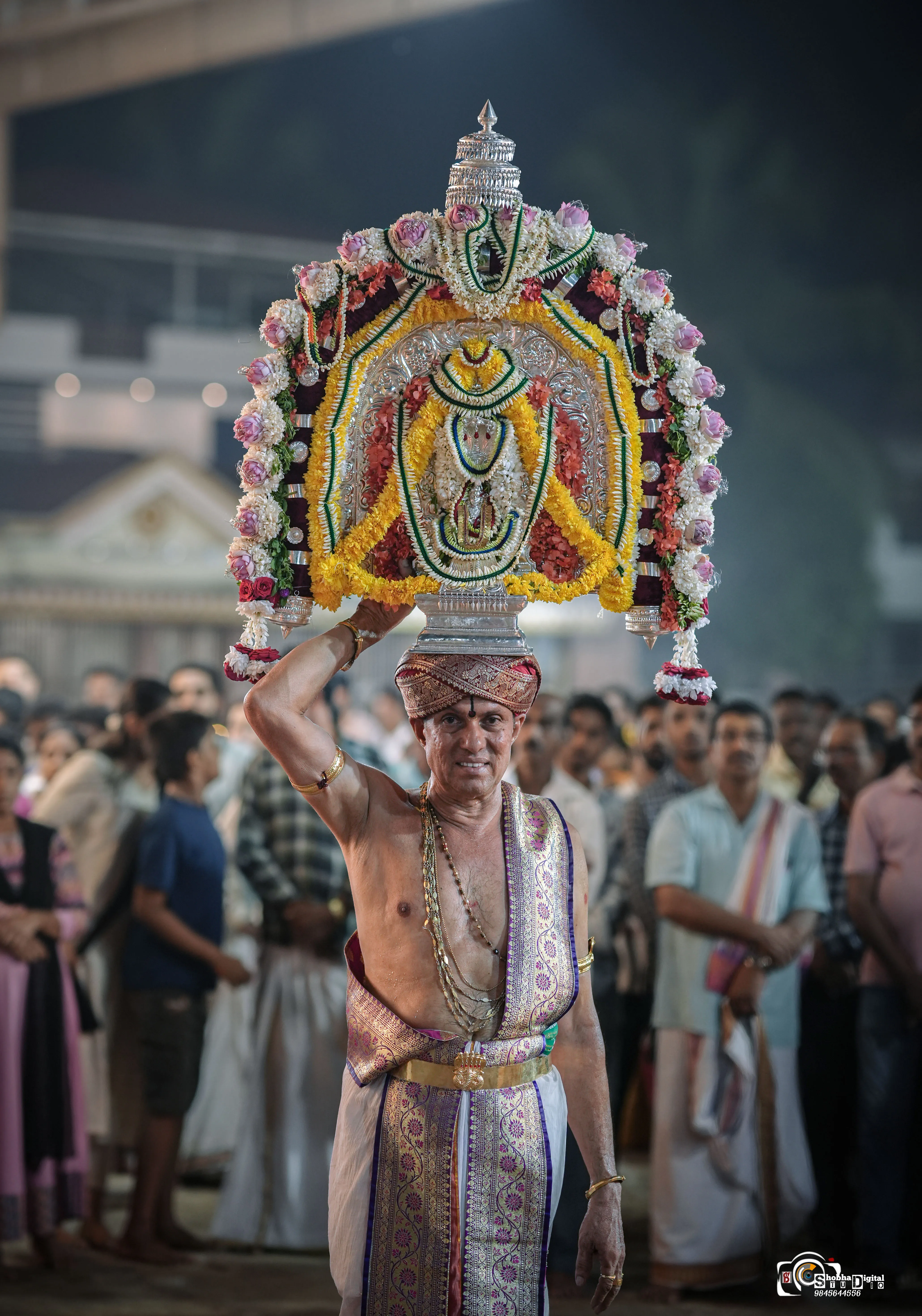 A devotee carrying an ornate deity crown at a temple festival, photographed by Shobha Digital Studio