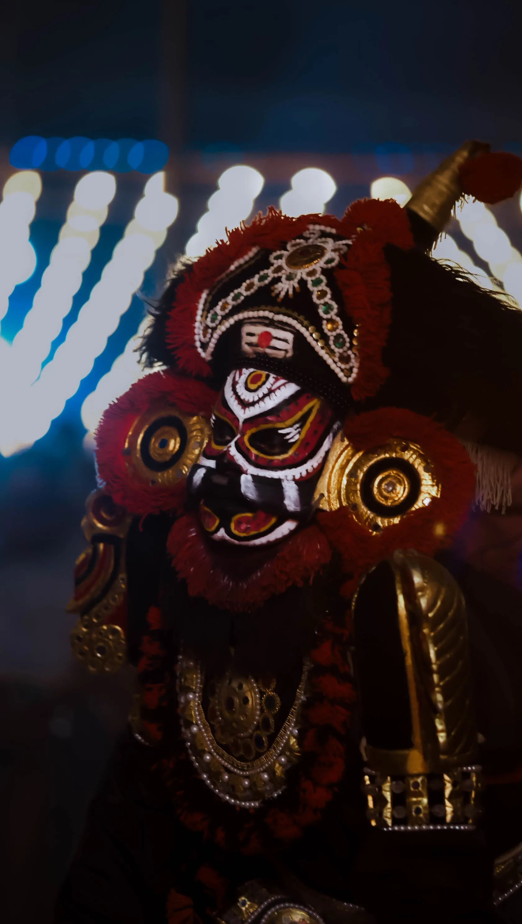 Yakshagana performer in fierce red, white and black demon face paint and a jeweled crimson crown, lit against glowing blue festival lights at night